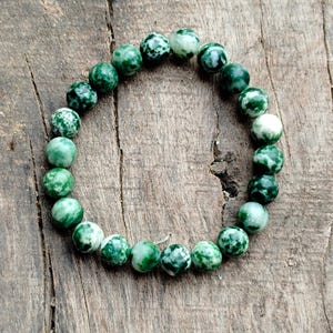 Könnte beinhalten: Ein Armband mit runden, grün-weiß gesprenkelten Perlen. Die Perlen sind zu einem Armband aufgereiht. Die Perlen haben einen Durchmesser von etwa 0,8 cm. Das Armband liegt auf einer Holzoberfläche.