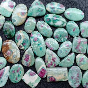 Peut inclure: Une collection de cabochons de pierres précieuses vertes et roses. Les pierres sont de forme ovale, ronde et rectangulaire. Elles présentent un motif vert et rose marbré. Les pierres sont disposées de manière aléatoire sur une surface noire. @TheGemsAndJewelsIn