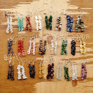 Op de afbeelding: Een verzameling edelsteen oorbellen in diverse kleuren, waaronder wit, blauw, rood, groen en zwart. Elke oorbel heeft een waterval van kleine, onregelmatig gevormde stenen die aan een zilveren haak hangen.