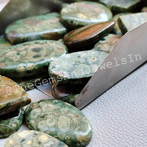 May include: A collection of green, brown, and white oval-shaped polished stones. The stones have a unique pattern and are arranged in a metal tray.