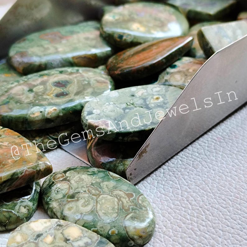 May include: A collection of green, brown, and white oval-shaped polished stones. The stones have a unique pattern and are arranged in a metal tray.