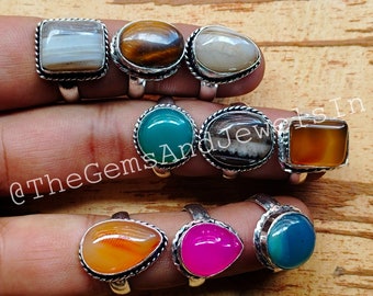 May include: A collection of nine silver rings with different colored gemstone inlays. The rings feature a variety of shapes and sizes, including round, oval, and square. The gemstones are set in silver bezels and have a polished finish. The rings are displayed on a hand.