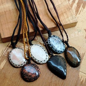 Op de afbeelding: Zes verschillende edelsteen hangers aan zwarte koorden. De hangers zijn allemaal ovaal of druppelvormig en hebben een verscheidenheid aan kleuren en patronen. De hangers zijn allemaal omgeven door een zwart koord dat bovenaan in een knoop is geknoopt.