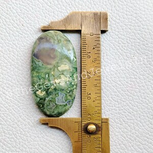 May include: A green, brown, and white oval-shaped cabochon gemstone measuring approximately 1.5 inches in length. The gemstone is being measured with a ruler.