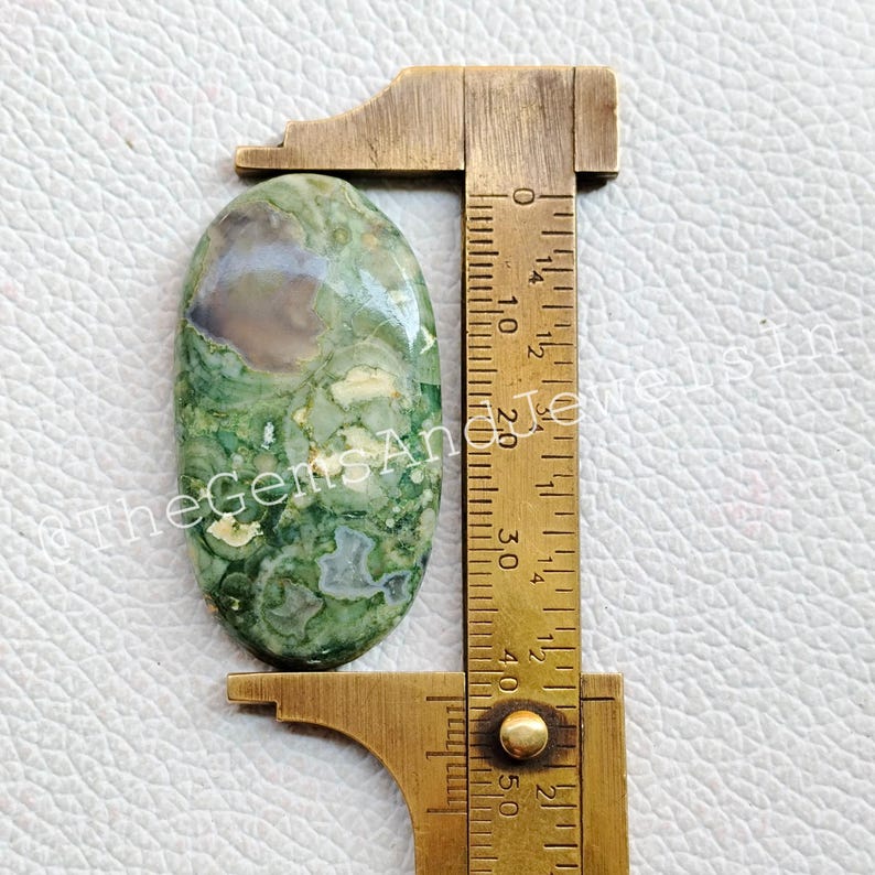 May include: A green, brown, and white oval-shaped cabochon gemstone measuring approximately 1.5 inches in length. The gemstone is being measured with a ruler.