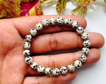 May include: A beaded bracelet featuring round, cream-colored beads with black spots, reminiscent of a Dalmatian dog's coat. The bracelet is strung together, creating a circular shape. The beads are approximately 0.3 inches in diameter.
