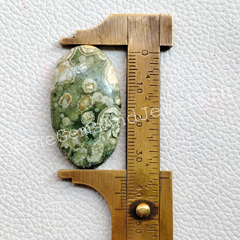 May include: A green, brown, and white oval-shaped ocean jasper cabochon measuring approximately 2.5 cm in length. The cabochon is being measured with a ruler.