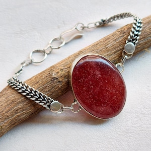 May include: A silver bracelet with a large, oval, red gemstone. The gemstone is set in a silver bezel and attached to a chain-link bracelet. The bracelet is displayed on a piece of wood.