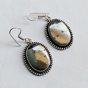 Può includere: Orecchini color argento con pietre ovali in pirite. Le pietre hanno un aspetto metallico oro e argento, incastonate in un bordo di perline. Chiusura a gancio.