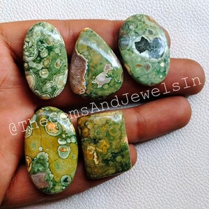 May include: Six green, brown, and white polished gemstone cabochons with a variety of patterns and shapes. The cabochons are displayed on a hand.