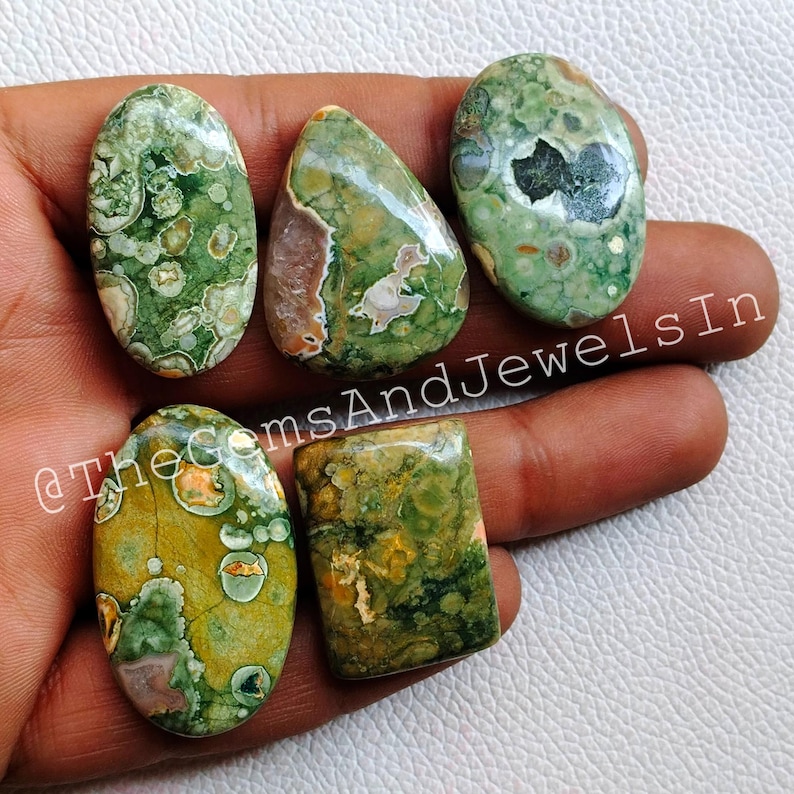 May include: Six green, brown, and white polished gemstone cabochons with a variety of patterns and shapes. The cabochons are displayed on a hand.