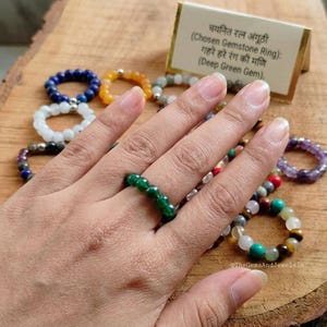Puede incluir: Una mano que lleva un anillo de piedra preciosa verde oscuro, rodeada de varios anillos de cuentas de diferentes colores. Los anillos se exhiben sobre una superficie de madera. Se ve una pequeña tarjeta con texto en inglés y otro idioma.
