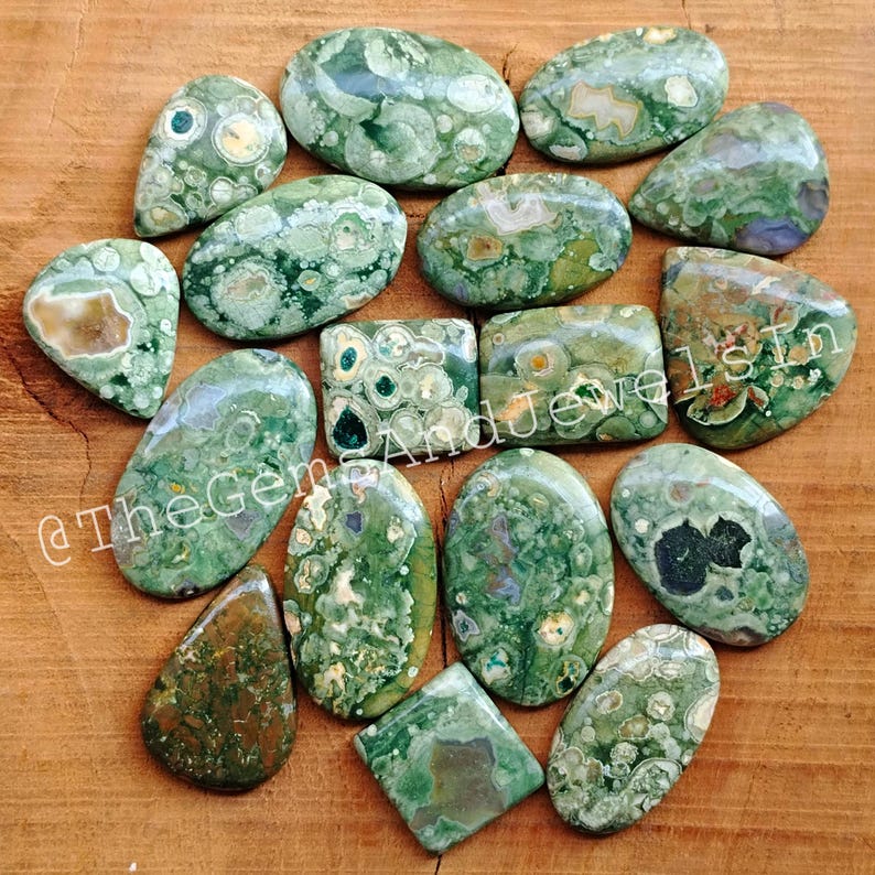 May include: A collection of 16 green, brown, and white ocean jasper cabochons. The cabochons are various shapes and sizes, including oval, teardrop, and square.
