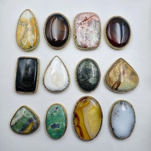 Könnte beinhalten: Eine Sammlung von zwölf Cabochon-Edelsteinen, die in goldfarbene, seilartig texturierte Metallfassungen eingefasst sind. Die Steine haben verschiedene Formen wie oval, birnenförmig und rechteckig und zeigen diverse Muster und Farben.