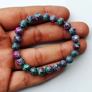 May include: A bracelet made of round gemstones in teal, magenta, and grey. The gemstones are approximately 0.8 cm in diameter. The bracelet is strung on an elastic cord. This is a piece of jewellery.