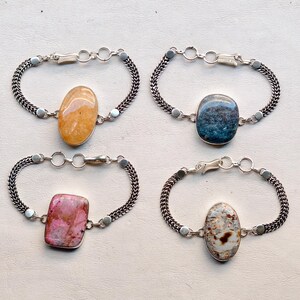May include: Four silver bracelets with different gemstone centerpieces. The gemstones are oval or rectangular, in shades of orange, blue, pink, and white with brown speckles. The bracelets have a chain-like design and a clasp closure.