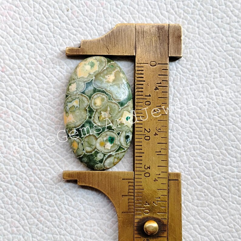 May include: Oval shaped green, white, and brown jasper cabochon measuring approximately 2.5 cm in length. The cabochon is being measured with a ruler.