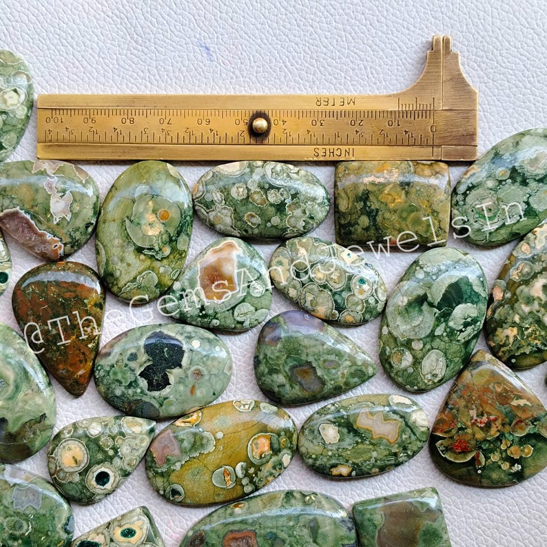 May include: A collection of 20 green, brown, and white ocean jasper cabochons. The cabochons are various shapes and sizes, with a speckled pattern. The cabochons are arranged on a white surface with a ruler for scale.