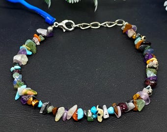 Op de afbeelding: Een kleurrijke armband met een verscheidenheid aan kleine, onregelmatig gevormde edelstenen in tinten blauw, groen, paars, oranje en bruin. De armband is geregen op een zilverkleurige ketting en heeft een sluiting. De edelstenen zijn ongeveer 0,6 cm groot.