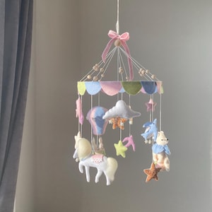 Puede incluir: Móvil para bebé en tonos pastel, con figuras de fieltro de un caballo, globo aerostático, nubes, estrellas y un oso. El móvil está suspendido de una cinta rosa y cuentas de madera. El diseño presenta colores suaves como el rosa, azul y amarillo.