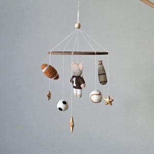 May include: A baby mobile featuring a brown wooden ring with various sports-themed felt ornaments. Includes a football, baseball, soccer ball, a teddy bear wearing a jersey with the number 1, a baseball bat, and star-shaped accents.