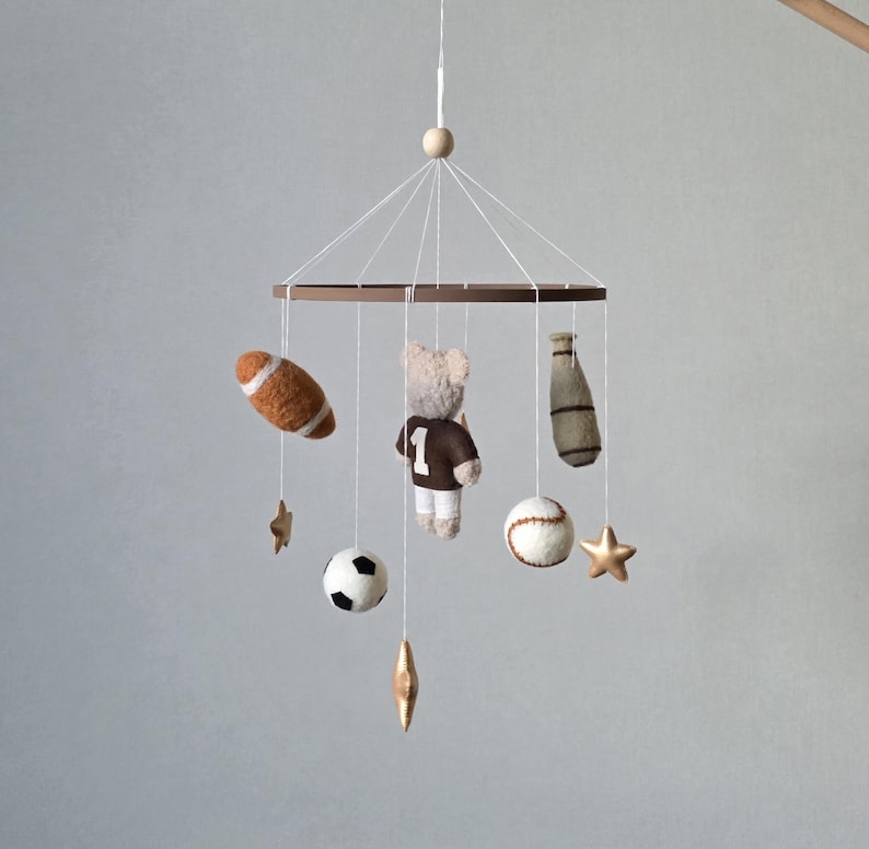 May include: A baby mobile featuring a brown wooden ring with various sports-themed felt ornaments. Includes a football, baseball, soccer ball, a teddy bear wearing a jersey with the number 1, a baseball bat, and star-shaped accents.
