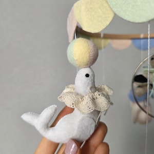 May include: A handmade baby mobile featuring a white felt seal balancing a colorful ball on its nose. The seal wears a lace collar. The mobile has pastel-colored felt accents and a wooden frame, creating a whimsical nursery decoration.