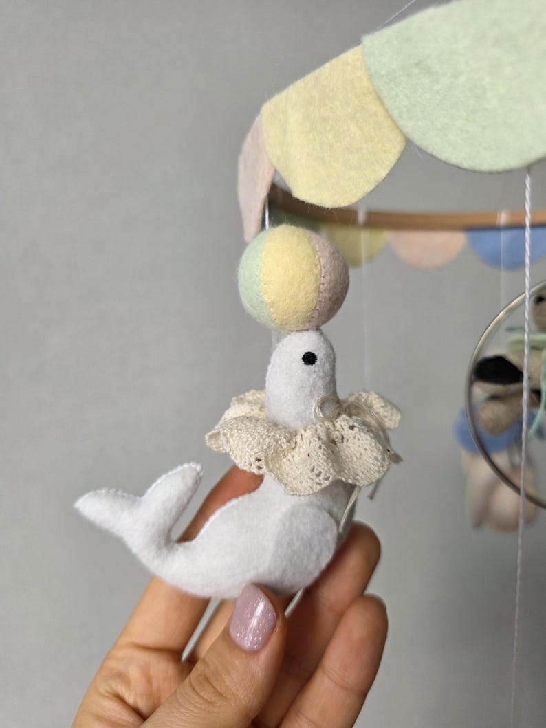 May include: A handmade baby mobile featuring a white felt seal balancing a colorful ball on its nose. The seal wears a lace collar. The mobile has pastel-colored felt accents and a wooden frame, creating a whimsical nursery decoration.