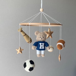 May include: A baby mobile featuring a teddy bear wearing a blue shirt with a white "H", alongside sports-themed felt ornaments. Includes a baseball bat, football, soccer ball, baseball, and gold stars, all suspended from a wooden hoop.