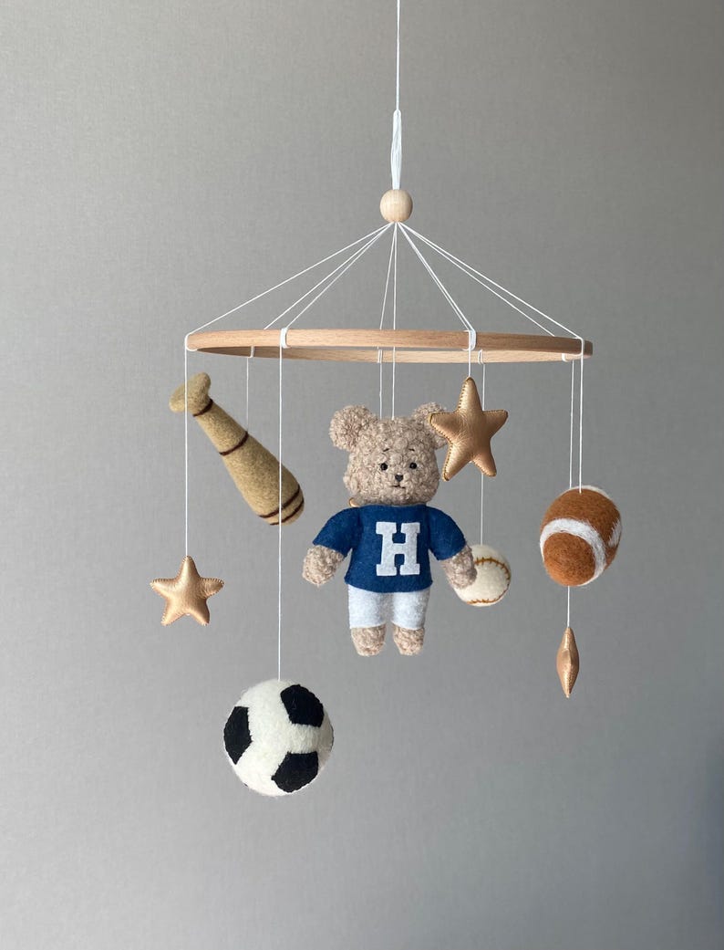 May include: A baby mobile featuring a teddy bear wearing a blue shirt with a white "H", alongside sports-themed felt ornaments. Includes a baseball bat, football, soccer ball, baseball, and gold stars, all suspended from a wooden hoop.
