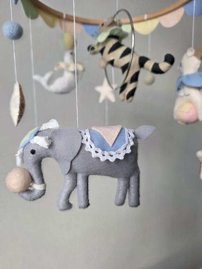 May include: A handmade baby mobile featuring felt circus animals. The mobile includes a gray elephant with a ball, a zebra in a hoop, a white seal, and other colorful felt shapes. The mobile is suspended from a wooden hoop.