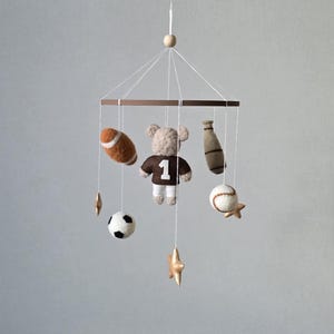 May include: A baby mobile featuring sports-themed felt ornaments. Includes a football, baseball, soccer ball, baseball bat, and a teddy bear wearing a jersey with the number 1. The mobile is suspended from a wooden ring.
