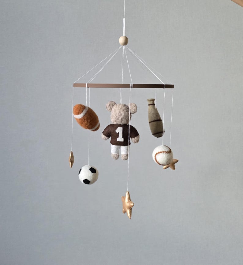 May include: A baby mobile featuring sports-themed felt ornaments. Includes a football, baseball, soccer ball, baseball bat, and a teddy bear wearing a jersey with the number 1. The mobile is suspended from a wooden ring.