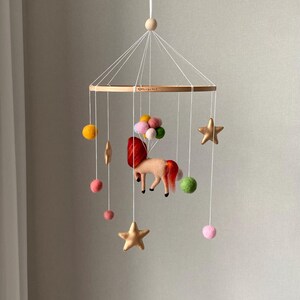 K&ouml;nnte beinhalten: Baby-Mobile mit einem Filz-Einhorn, roter M&auml;hne und Schweif, das an einem Holzreifen h&auml;ngt. Es hat goldene Sterne, bunte Filzkugeln und eine Ansammlung von rosa und gr&uuml;nen Filzkugeln.