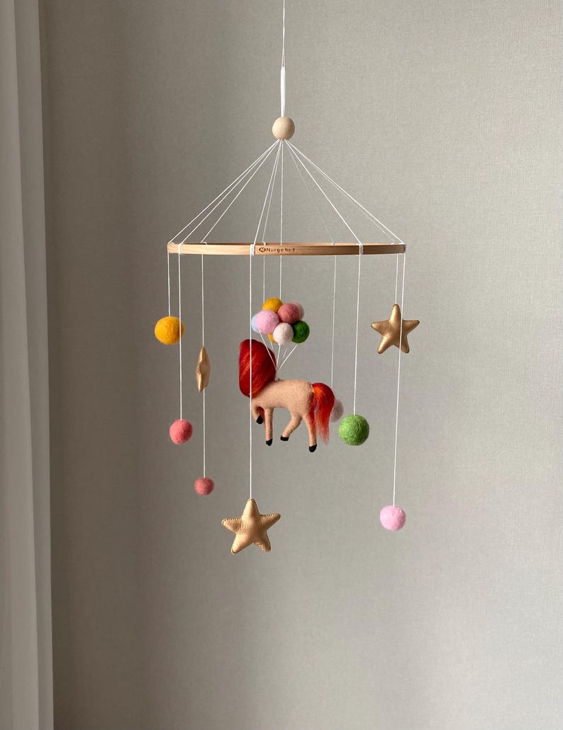 K&ouml;nnte beinhalten: Baby-Mobile mit einem Filz-Einhorn, roter M&auml;hne und Schweif, das an einem Holzreifen h&auml;ngt. Es hat goldene Sterne, bunte Filzkugeln und eine Ansammlung von rosa und gr&uuml;nen Filzkugeln.