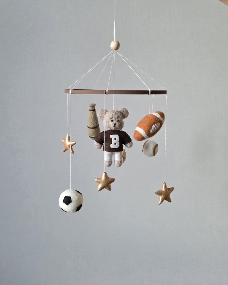 May include: A baby mobile featuring a teddy bear wearing a brown shirt with a white "B", a football, baseball, soccer ball, a bottle, and gold stars. The mobile is suspended from a wooden ring and a wooden bead.