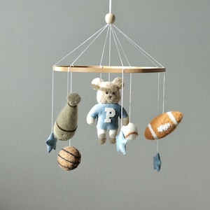 May include: A baby mobile featuring a wooden ring with hanging felt toys. The toys include a teddy bear in a blue sweater, a football, a baseball, a basketball, a bottle, and blue star shapes. The mobile is suspended by white strings.
