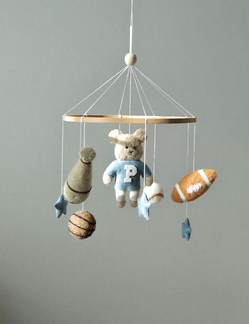 May include: A baby mobile featuring a wooden ring with hanging felt toys. The toys include a teddy bear in a blue sweater, a football, a baseball, a basketball, a bottle, and blue star shapes. The mobile is suspended by white strings.