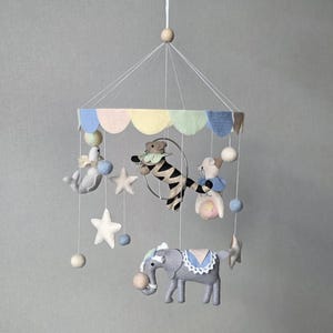 May include: A whimsical baby mobile featuring felt animal figures, including a gray elephant, a zebra, and a seal. The mobile has a carousel design with pastel-colored accents, stars, and hanging felt balls. The mobile is suspended from a wooden bead.