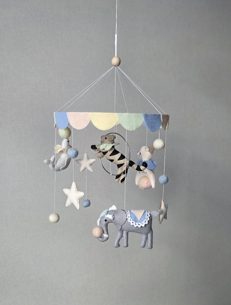 May include: A whimsical baby mobile featuring felt animal figures, including a gray elephant, a zebra, and a seal. The mobile has a carousel design with pastel-colored accents, stars, and hanging felt balls. The mobile is suspended from a wooden bead.