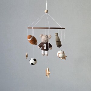 May include: A baby mobile featuring sports-themed felt ornaments. Includes a football, baseball, soccer ball, baseball bat, and a teddy bear wearing a jersey with the number 1. The mobile is suspended from a wooden ring with gold star accents.