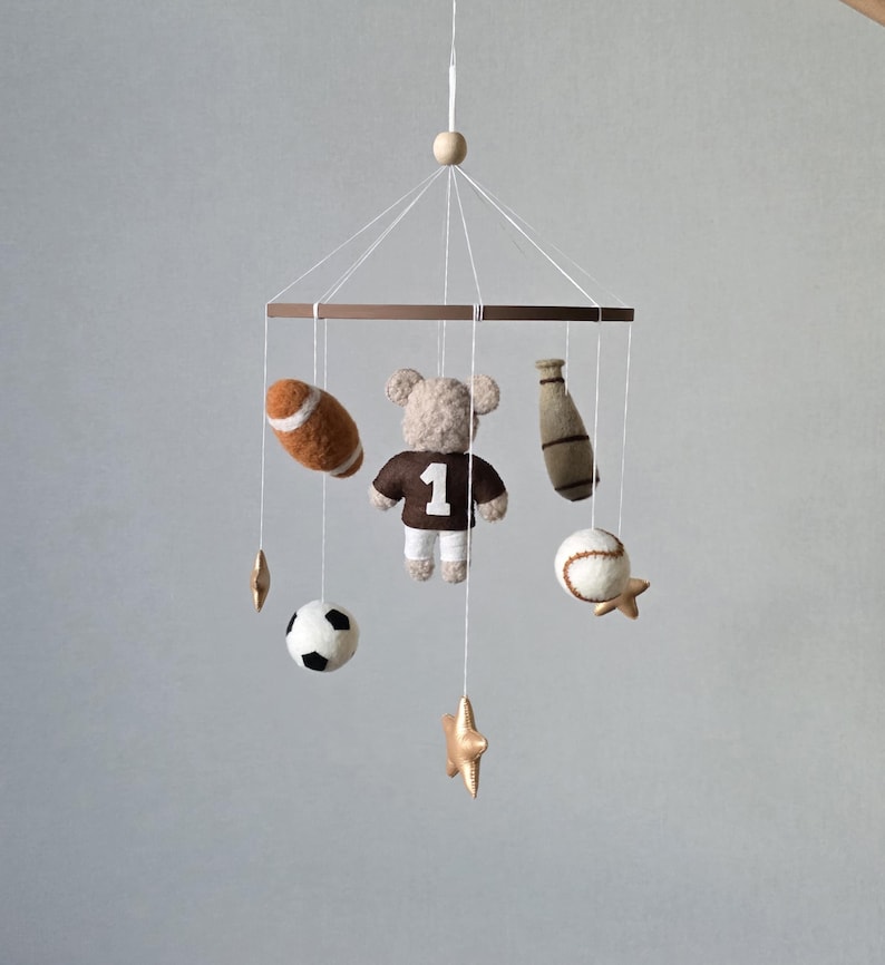 May include: A baby mobile featuring sports-themed felt ornaments. Includes a football, baseball, soccer ball, baseball bat, and a teddy bear wearing a jersey with the number 1. The mobile is suspended from a wooden ring with gold star accents.