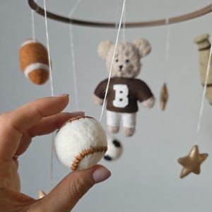 May include: A handmade baby mobile featuring felt sports-themed elements. Includes a baseball, football, soccer ball, a teddy bear wearing a brown jersey with a white "B", and gold stars. The mobile is suspended from a brown wooden ring.