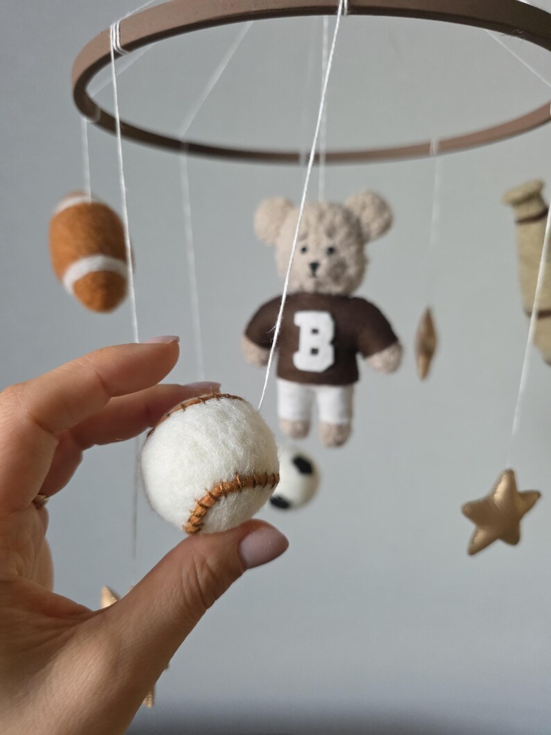 May include: A handmade baby mobile featuring felt sports-themed elements. Includes a baseball, football, soccer ball, a teddy bear wearing a brown jersey with a white "B", and gold stars. The mobile is suspended from a brown wooden ring.