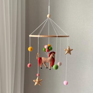 K&ouml;nnte beinhalten: Baby-Mobile mit Holzring und h&auml;ngenden Filzdekorationen. Das Mobile zeigt ein beiges Pferd mit roter M&auml;hne, goldene Sterne, bunte Filzb&auml;lle und Pompons. Es h&auml;ngt an einer wei&szlig;en Schnur. Ma&szlig;e in Zentimetern.