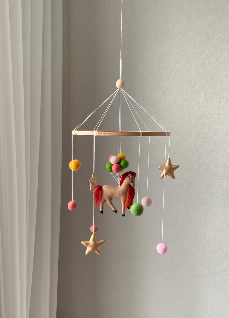 K&ouml;nnte beinhalten: Baby-Mobile mit Holzring und h&auml;ngenden Filzdekorationen. Das Mobile zeigt ein beiges Pferd mit roter M&auml;hne, goldene Sterne, bunte Filzb&auml;lle und Pompons. Es h&auml;ngt an einer wei&szlig;en Schnur. Ma&szlig;e in Zentimetern.
