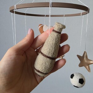 May include: A handmade baby mobile featuring a brown wooden ring with hanging felt shapes. The shapes include a football, a baseball bat, a soccer ball, and a gold star. The mobile is suspended by white strings.