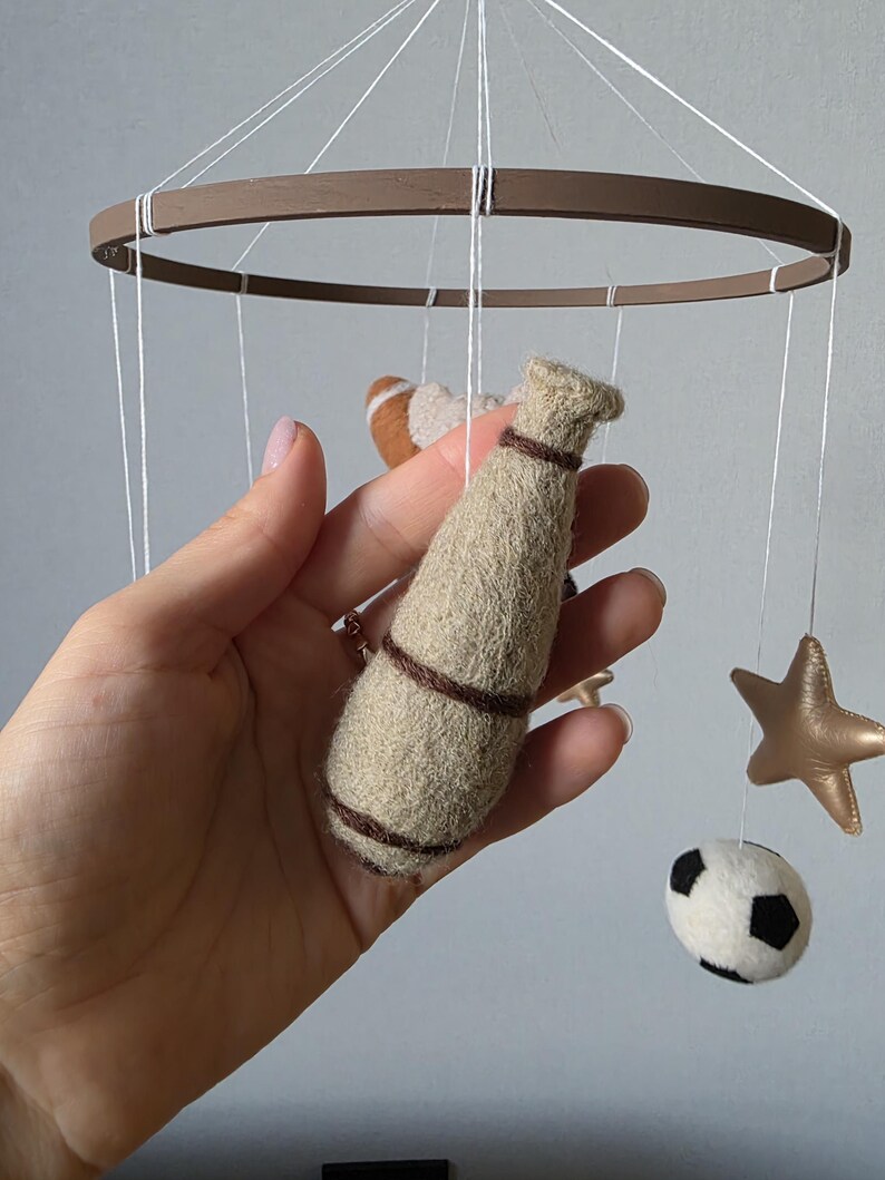 May include: A handmade baby mobile featuring a brown wooden ring with hanging felt shapes. The shapes include a football, a baseball bat, a soccer ball, and a gold star. The mobile is suspended by white strings.
