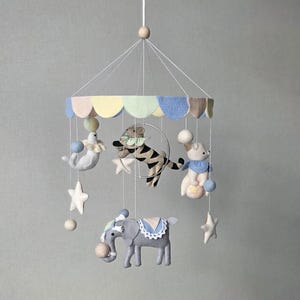 May include: A whimsical baby mobile featuring felt circus animals, including a tiger, seal, bear, and elephant. The mobile has a pastel-colored canopy and is adorned with stars and pom-poms, creating a charming nursery decoration.