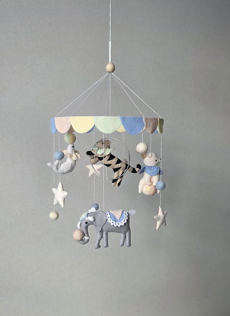 May include: A whimsical baby mobile featuring felt circus animals, including a tiger, seal, bear, and elephant. The mobile has a pastel-colored canopy and is adorned with stars and pom-poms, creating a charming nursery decoration.
